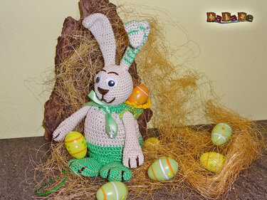 Gehäkelter Amigurumi-Osterhase in Beige und Grün mit kleinem Körbchen im Stroh, umgeben von bunten Ostereiern