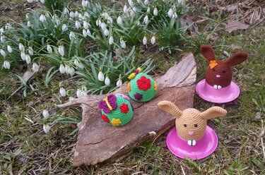 Verschiedene gehäkelte Eierwärmer mit Hasenohren und Blumenapplikationen auf Holz und Gras vor Schneeglöckchen