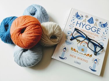 Fünf Knäuel Wolle in Blau-, Orange- und Cremetönen neben einem Buch und einer Brille.