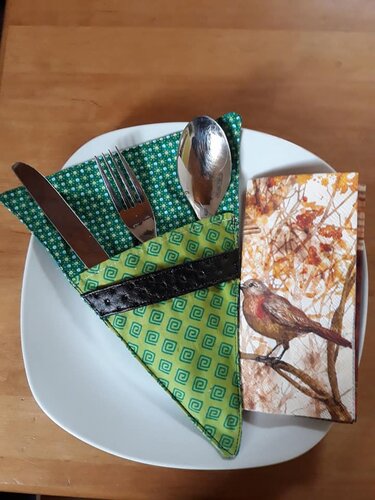 Dreieckige grüne Stoff-Bestecktasche mit Messer, Gabel und Löffel auf weißem Teller neben Serviette mit Vogelmotiv