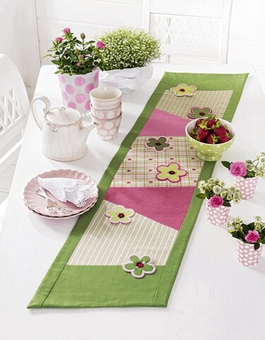 Patchwork-Tischläufer mit grünem Rand und aufgenähten Blumen in Grün, Rosa und Beige auf einem weißen Tisch