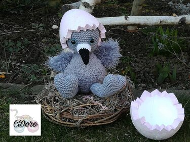 Grau gehäkeltes Küken-Amigurumi mit großen Augen und rosafarbener Eierschale im braunen Nest.