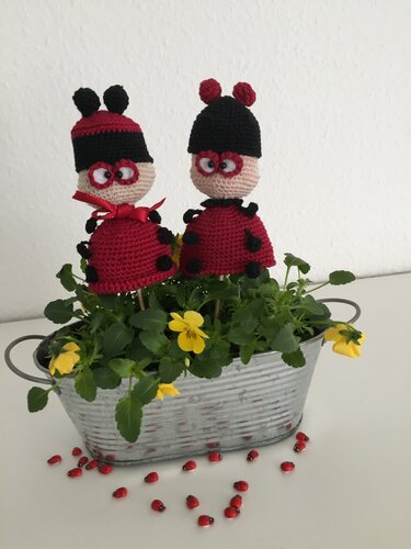 Zwei gehäkelte rote Marienkäfer-Figuren als Gartenstecker in einem ovalen Metallkübel mit gelben Stiefmütterchen und kleinen Marienkäfer-Dekoobjekten.