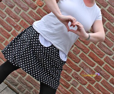 Person in schwarzem, weiß-getupftem Rock und hellem Oberteil formt mit den Händen ein Herz vor einer Ziegelwand.