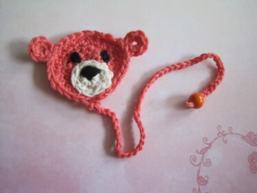 Nahaufnahme eines rosafarbenen gehäkelten Teddybär-Lesezeichens mit weißer Schnauze, schwarzen Augen und einer Schnur mit orangefarbener Perle.
