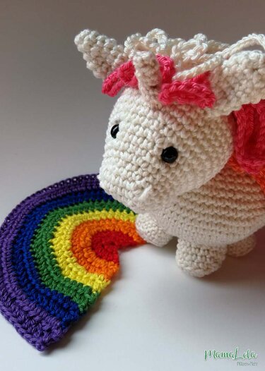 Weißes gehäkeltes Amigurumi-Einhorn mit schwarzen Augen und rosa Akzenten neben einem bunten gehäkelten Regenbogen.