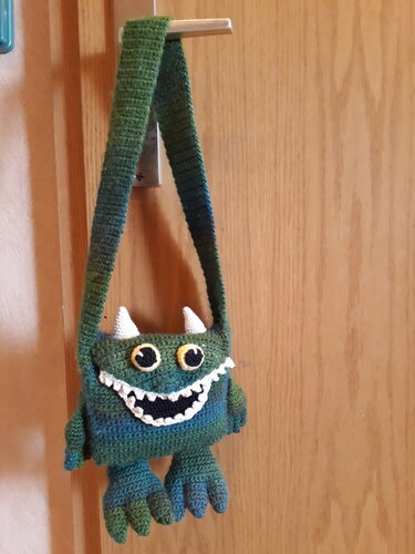 Gehäkelte Monster-Umhängetasche in Grün-Blau mit Hörnern, großen Augen und gezacktem Mund, an Türgriff hängend.