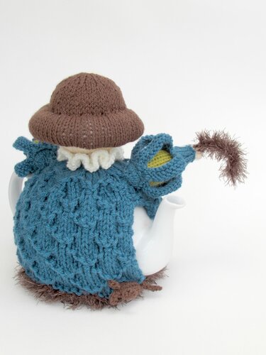 White teapot dressed in a whimsical blue knitted cozy with a brown hat and ruffled collar