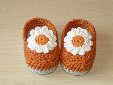 Nahaufnahme gehäkelter orangefarbener Babyschuhe mit weißer Blüte und hellblauer Sohle auf hellem Holz