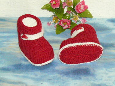 Nahaufnahme gestrickter roter Babyschuhe mit sichtbarer Strickstruktur, weißer Einfassung und Riemchen mit Pilzknopf