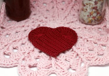 Handgehäkeltes rotes Herz auf einem hellrosa Häkeldeckchen mit detailreicher Musterung.