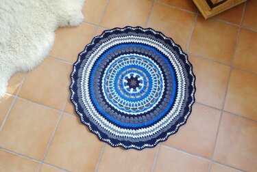 Runder gehäkelter Teppich mit Mandala‑Muster in Blau, Grau und Weiß auf terrakottafarbenem Fliesenboden