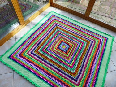 Quadratische, bunte Häkeldecke mit Granny-Square-Muster und hellgrünem Wellenrand auf Fliesenboden.