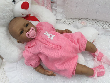 Baby im rosa gestrickten Strampler mit Hasen-Applikation und passenden gestrickten Babyschühchen mit Schleifen auf weißer Decke