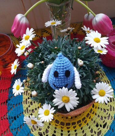 Blaues gehäkeltes Amigurumi in einem Topf mit weißen Gänseblümchen auf einem bunten Häkeldeckchen.