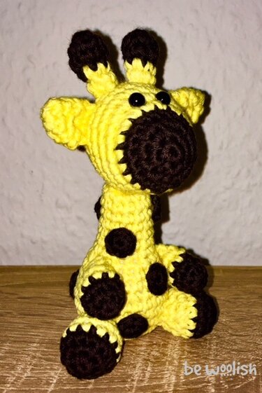 Gehäkelte sitzende Amigurumi-Giraffe in Gelb mit dunkelbraunen Flecken auf Holzuntergrund.