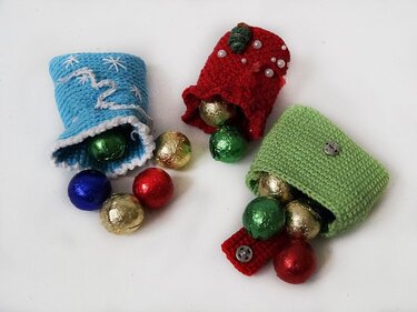 Drei gehäkelte Säckchen in Blau, Rot und Grün mit Schokokugeln auf weißem Hintergrund.