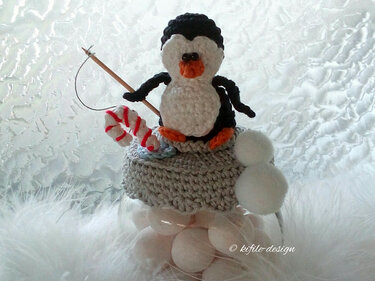 Gehäkelter schwarz-weißer Pinguin mit orangefarbenem Schnabel und Füßen auf grauem Häkeldeckel eines Glases, hält Angel mit rot-weißer Zuckerstange, daneben weiße Bommel.