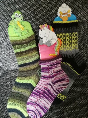 Drei handgestrickte Socken auf dunklem, strukturiertem Stoff: grün-braun gestreift, lila-rosa mit Lochmuster, grau-schwarz-gelb gemustert, mit Tier-Clips.