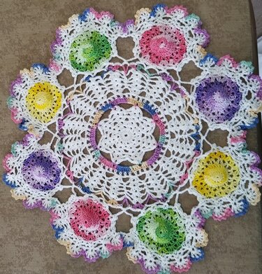 Round white crochet doily with vibrant multicolored floral centers and a pastel scalloped edge, on a brown surface.