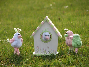 Gehäkelte Amigurumi-Vögel in Pastell rund um ein kleines Vogelhaus auf einer Wiese, ein Vogel sitzt im runden Fenster