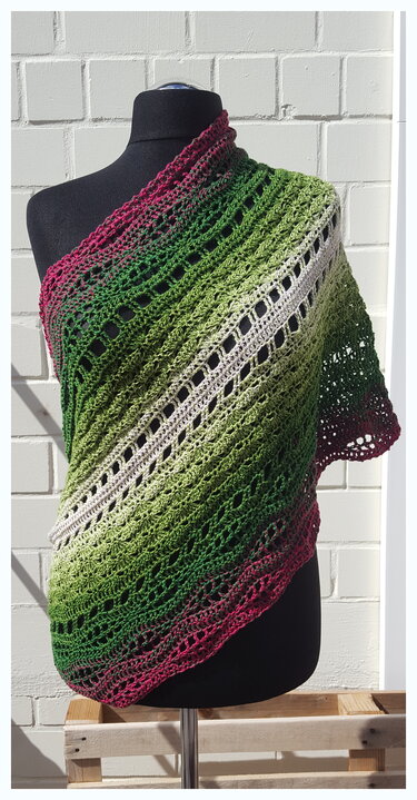 Gehäkeltes Schultertuch/Loop in Pink‑Grün mit diagonalem Lochmuster auf einer Schneiderpuppe vor heller Ziegelwand.