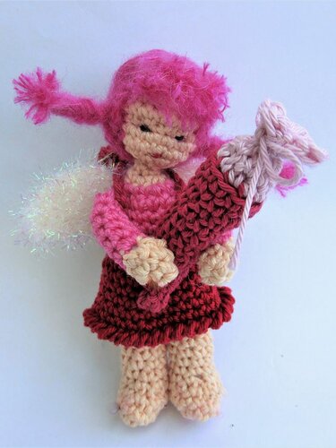 Gehäkeltes Amigurumi-Engelchen mit pinkem Haar, pink-rotem Kleid, cremefarbenen Beinen und glitzernden Flügeln, hält eine kleine Schultüte.