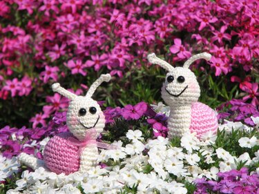 Zwei gehäkelte Schnecken mit rosa Häuschen zwischen weißen und pinken Blüten