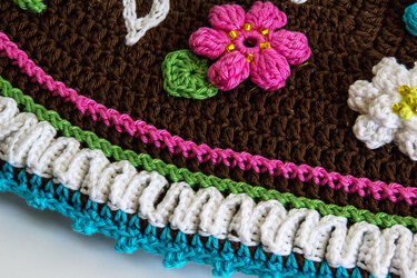 Nahaufnahme einer braunen Häkelarbeit mit pinkfarbener Perlenblume, grünem Blatt und mehrfarbigen Bordüren in Pink, Grün, Weiß und Türkis.