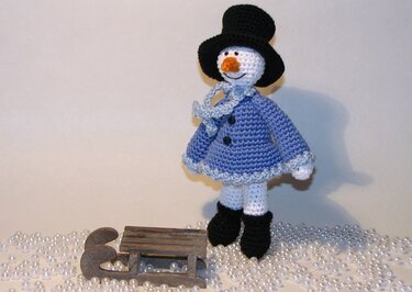 Gehäkelter Schneemann in blauer Jacke mit schwarzem Zylinder neben kleinem Holzschlitten auf Perlen