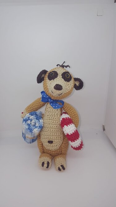 Gehäkeltes Amigurumi-Erdmännchen, braun-beige, mit blauer Schleife, blau-weißer Tasche und rot-weißem Objekt