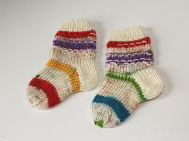 Zwei handgestrickte Babysocken in Creme mit bunten Streifen in Rot, Orange, Lila, Grün und Blau auf hellem Hintergrund.
