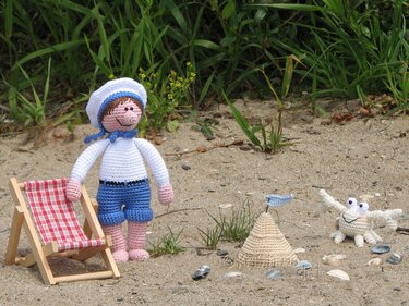 Gehäkelte Matrosenpuppe neben Liegestuhl, Sandburg und Amigurumi-Krabbe auf Sand