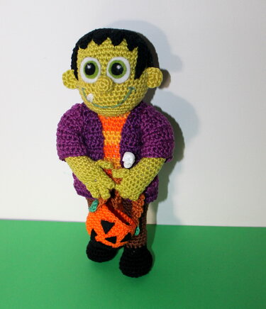 Green crochet amigurumi doll in purple jacket holding a pumpkin bag