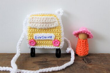 Gehäkelte gelb-weiße Bus-Tasche mit dem Schriftzug „SUNSHINE“ und einer kleinen Holzpuppe mit orangefarbenem Häkelkleid und pinkem Hut