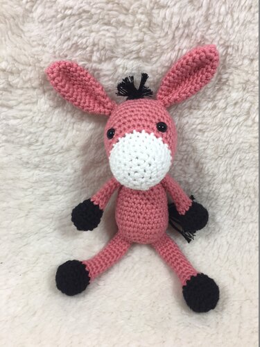 Rosa gehäkelter Amigurumi‑Esel mit weißer Schnauze und schwarzen Details, sitzend auf hellem, flauschigem Stoff.