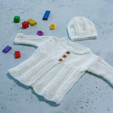Weiße, gestrickte Baby-Strickjacke mit drei orangefarbenen Blumenknöpfen und eine passende Strickmütze. Das Set liegt auf hellgrauem Untergrund, umgeben von bunten Buchstaben und Holzbausteinen.