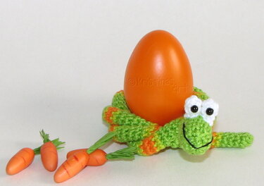 Gehäkelte grüne Tierfigur mit weißen Augen und schwarzem Lächeln, die ein orangefarbenes Ei hält; kleine Dekokarotten liegen daneben auf hellem Hintergrund.