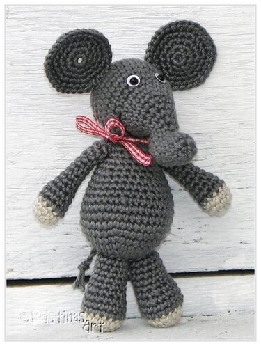 Gehäkelter grauer Amigurumi‑Elefant mit Kulleraugen, kariertem Schleifchen und hellen Pfoten auf hellem Holz.