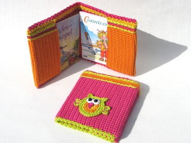 Pinkfarbene gehäkelte Buchhülle mit Eulen-Applikation vor aufgeklappter orangefarbener Hülle