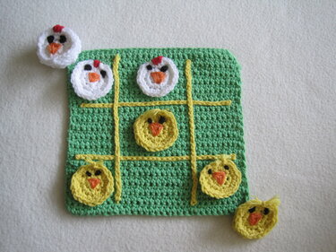 Grünes gehäkeltes Tic‑Tac‑Toe‑Brett mit gelbem Gitter und runden Küken- und Hühner-Spielsteinen