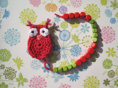 Mini gehäkelte rote Eule mit weißen Augen neben einer halbkreisförmigen Kette roter und grüner Perlen auf floralem Papier