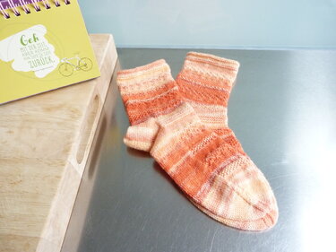 Handgestrickte Kurzsocken mit orange-cremefarbenem Farbverlauf auf Metallfläche neben Holzbrett.