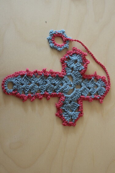 handgehäkeltes Kreuz in hellgrau mit rosa Umrandung und kleiner Ringöse, liegt auf hellem Holz