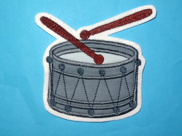 Bestickte graue Trommel mit zwei roten Schlägeln und weißer Umrandung auf blauem Hintergrund.