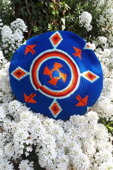 Runde blaue gehäkelte Umhängetasche mit orange‑rotem Ethno‑Muster auf weißen Blüten