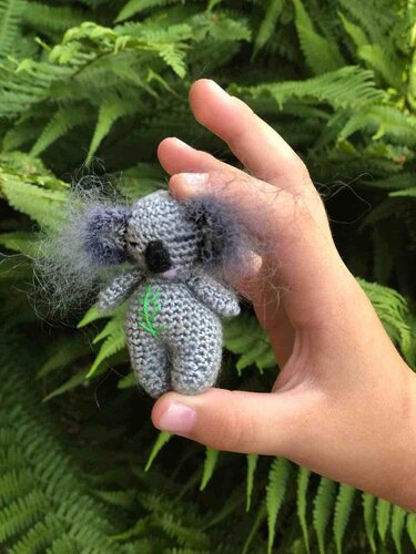 Hand hält winziges graues gehäkeltes Koala-Amigurumi vor Farnblättern.