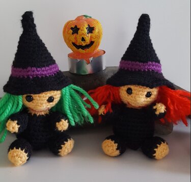 Zwei gehäkelte Amigurumi-Hexen mit grünen und roten Haaren, schwarzen Hüten und einem Kürbis-Teelicht
