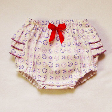 White baby bloomers with purple circle print, ruffled legs, and red bow