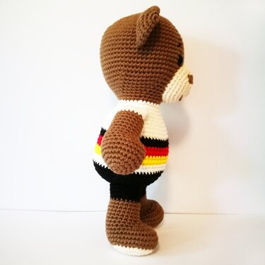 Gehäkelter brauner Amigurumi-Teddybär, Seitenansicht, weißes Shirt mit schwarzen, roten und gelben Streifen